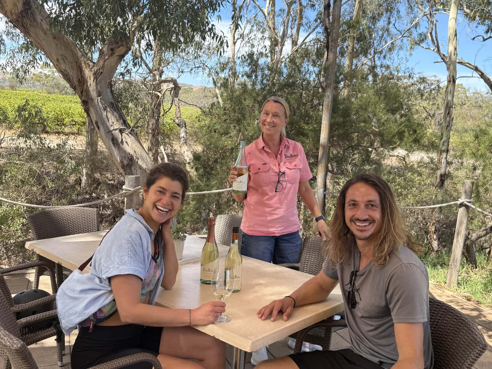 A woman serves wine to a young couple who are seated by river gums at DogRidge Cellar Door, McLaren Vale