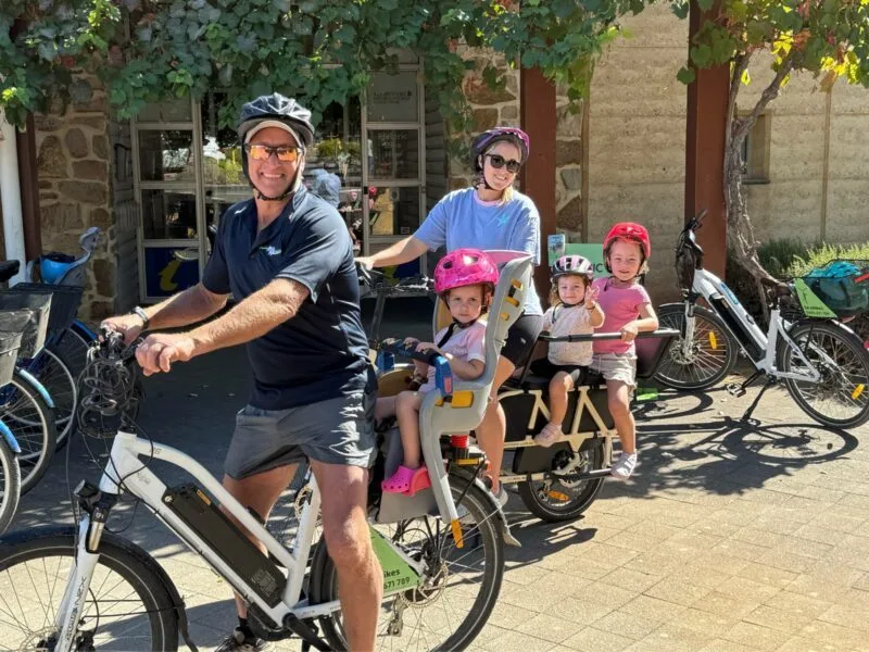 A man astride an e-bike with a small child in the child seat behind, and a woman astride a cargo bike with 2 young children on the bench seat behind her.
