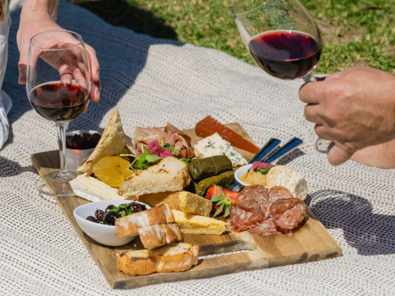 Glasses of red wine are held either side of a picnic platter