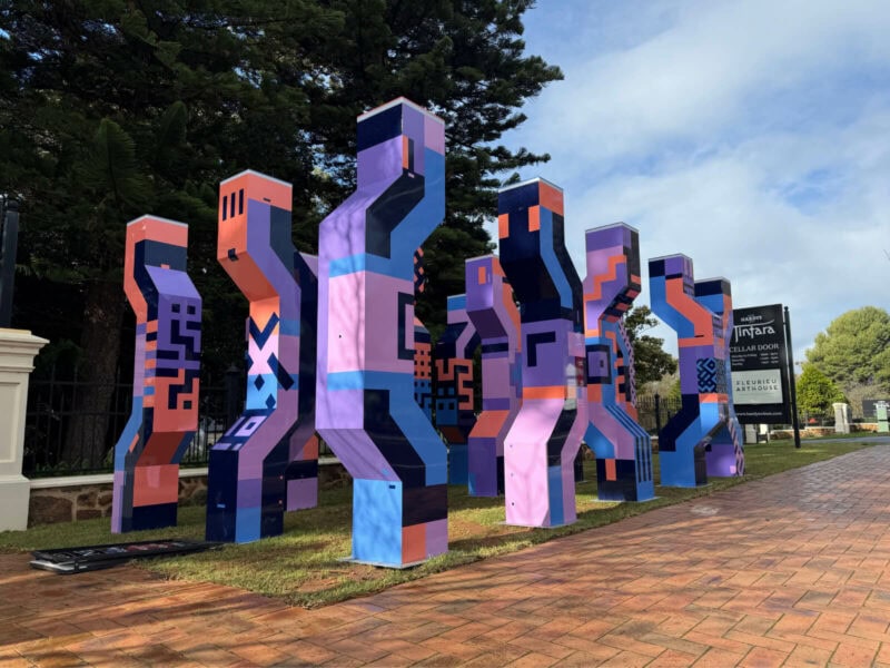 A sculptural installation of geometric multicoloured mettalic figures with the Hardys Tintara and tall pine trees in the background