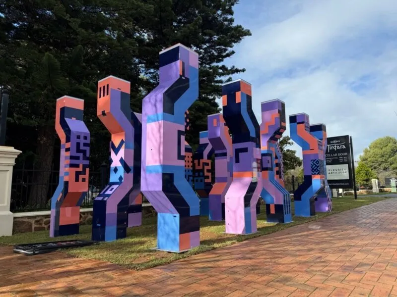 A sculptural installation of geometric multicoloured mettalic figures with the Hardys Tintara and tall pine trees in the background
