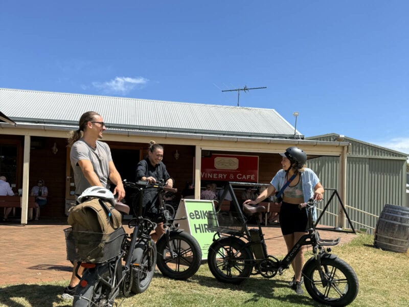Three young adults laughing as they lean on their eBikes in front of Oxenberry Farm Cafe, and SA eBikes signs.