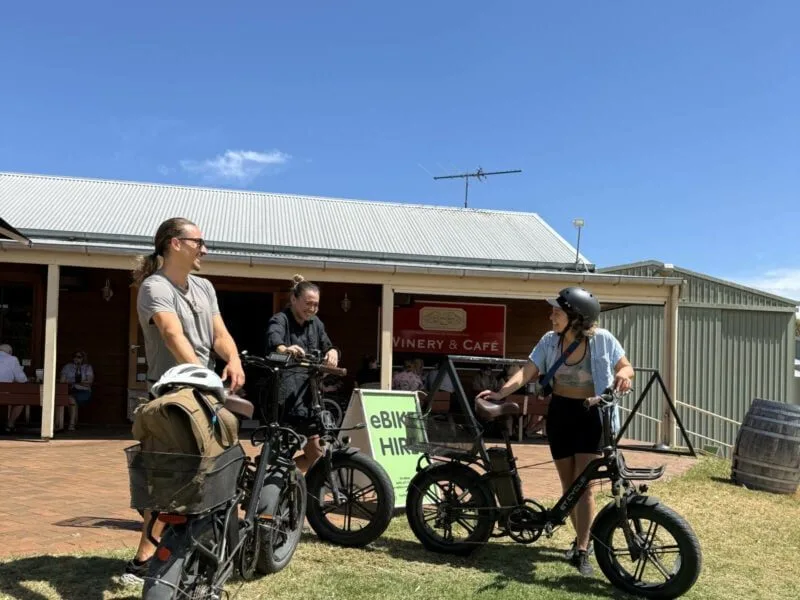 Three young adults laughing as they lean on their eBikes in front of Oxenberry Farm Cafe, and SA eBikes signs.