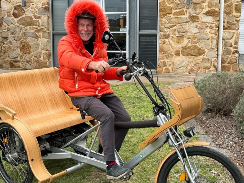 A man wearing an orange hooded jacket seated on the bike seat of an open top TukTuk