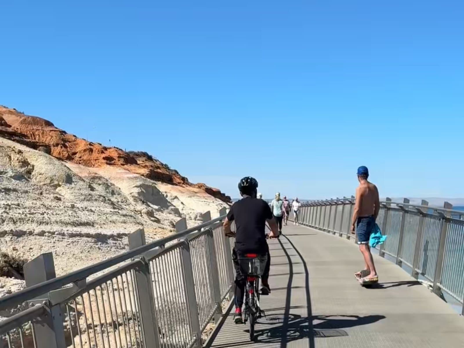 A cyclist and a skateboarder on the boardwalk, with the ochre hills of Witton Bluff, Port Noarlunga in the background