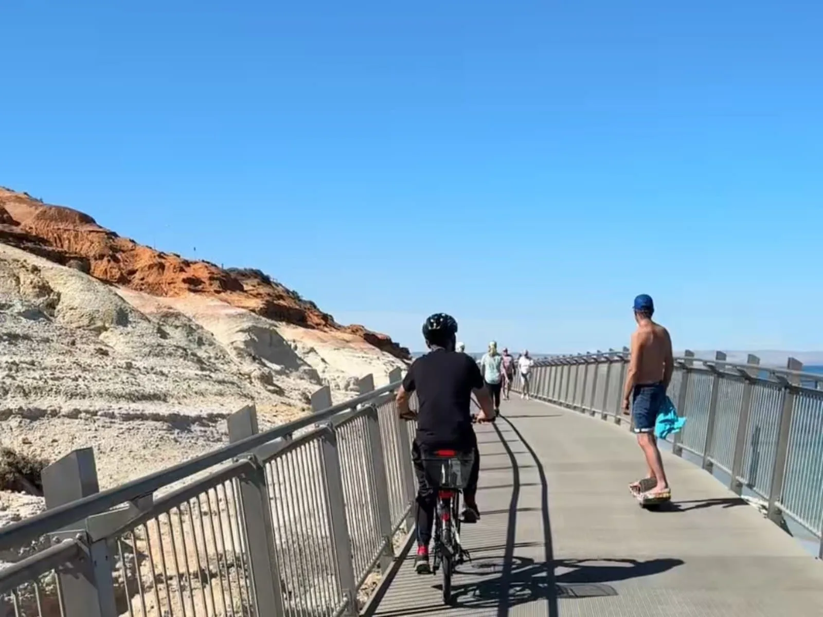 A cyclist and a skateboarder on the boardwalk, with the ochre hills of Witton Bluff, Port Noarlunga in the background