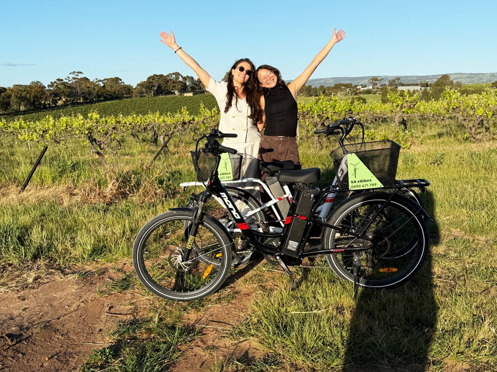 2 young women with arms raised in joy, standing behnd their ebikes