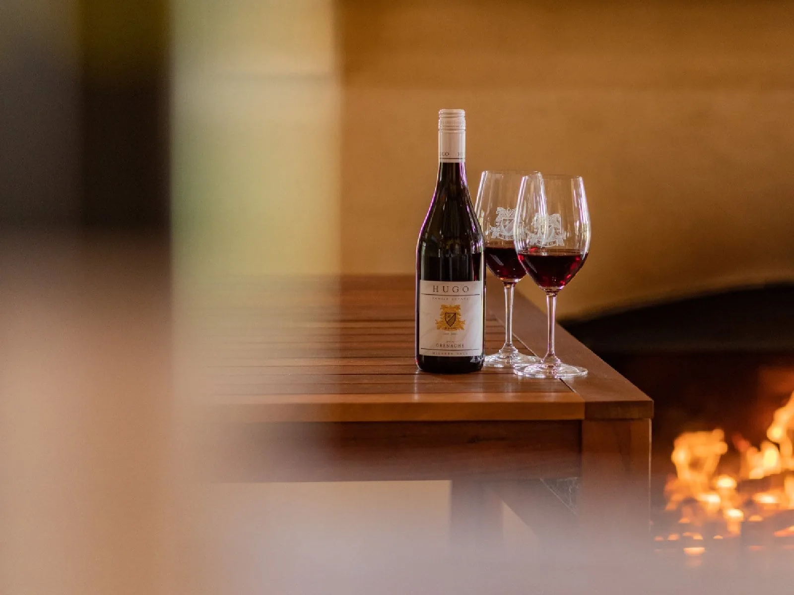 Bottle of Hugo Wine with 2 glasses of red wine and a fireplace in the background