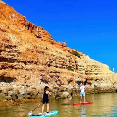 2 people standing up on paddle boards on calm seas with sandstone cliffs in the background