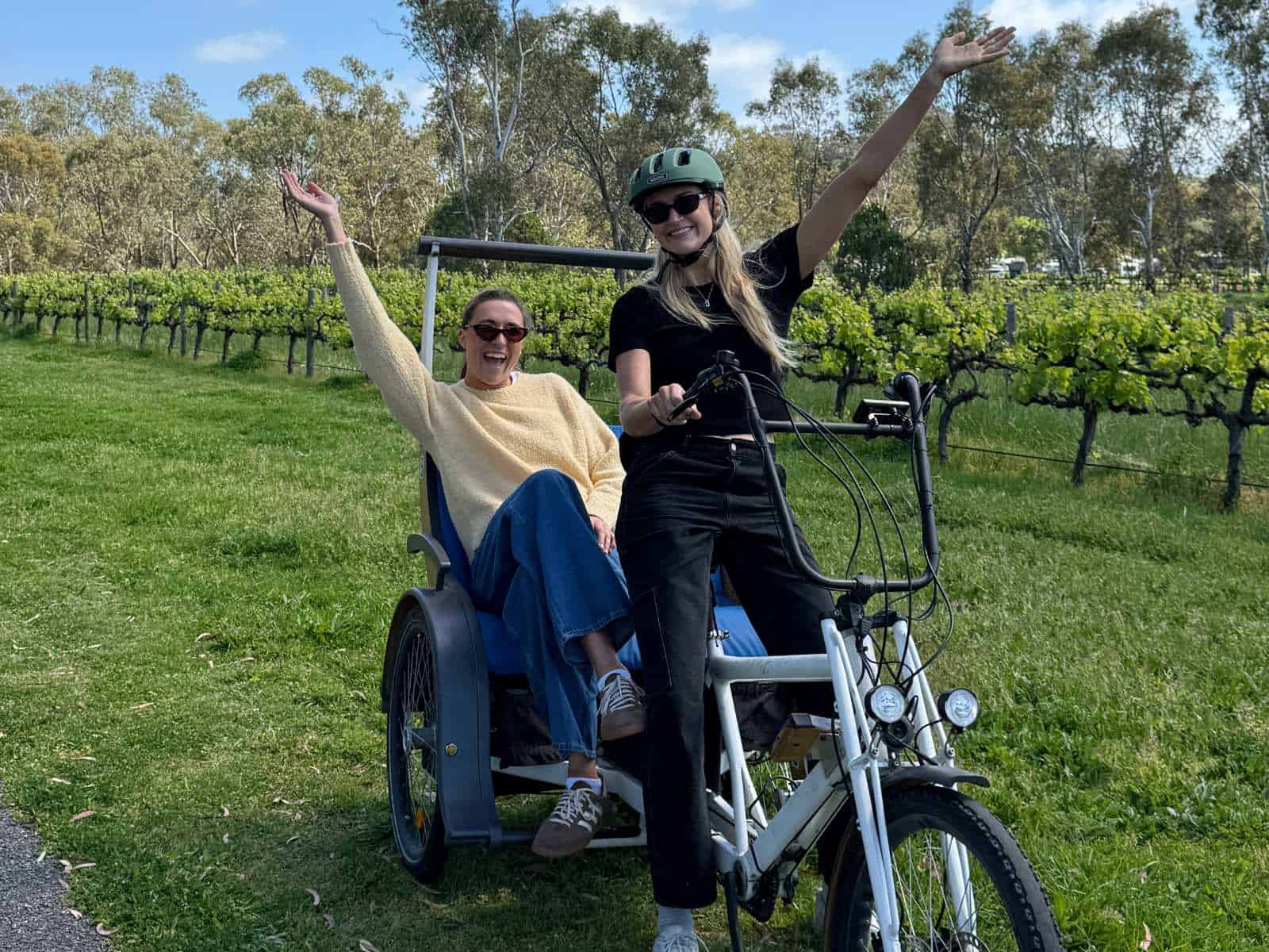 2 young women with arms raised whilst seated on an SA eBikes TukTuk in McLaren Vale with vines in the background